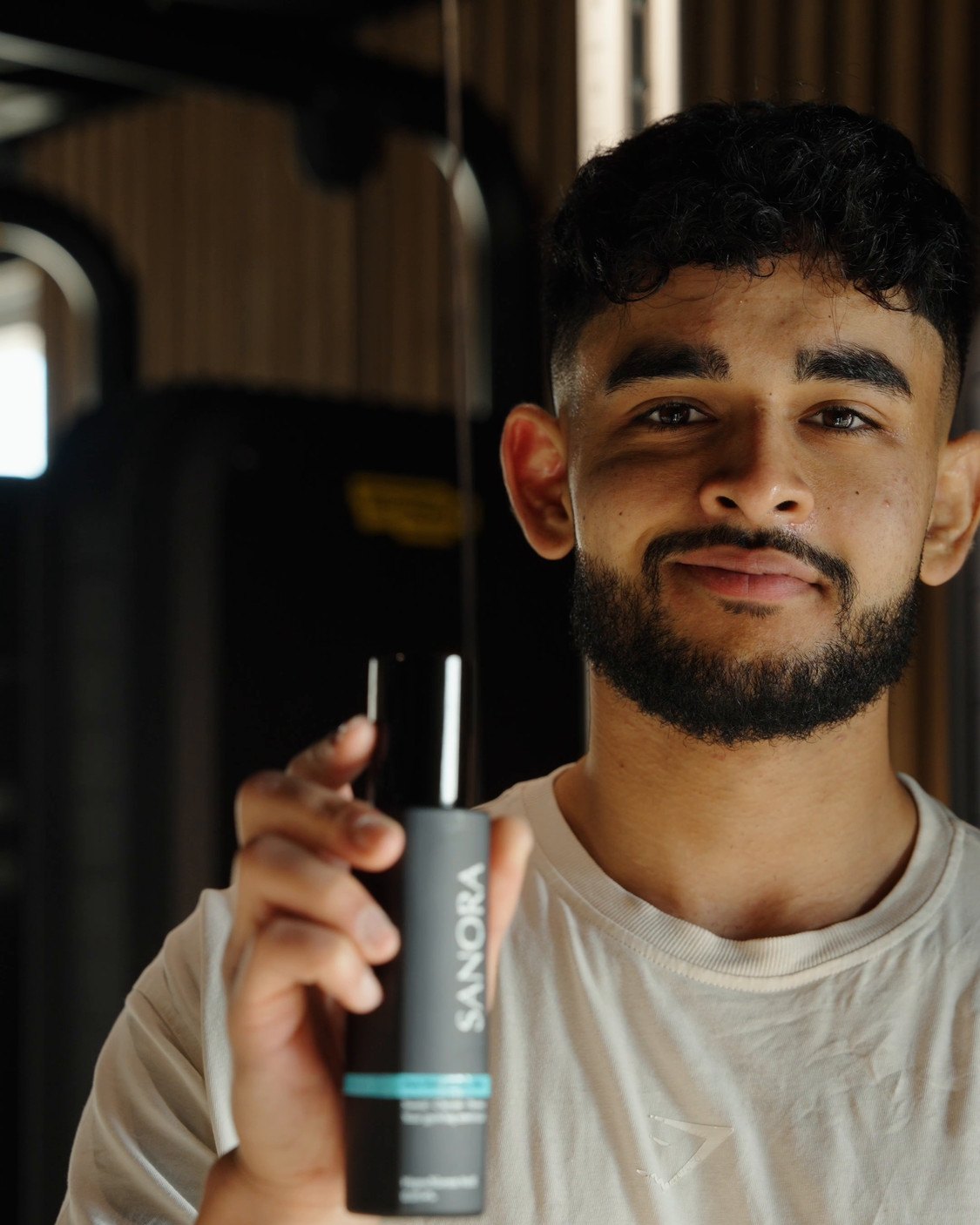 Harman, founder of Sanora, smiling and holding a bottle of Sanora Post-Workout Face Mist in the gym